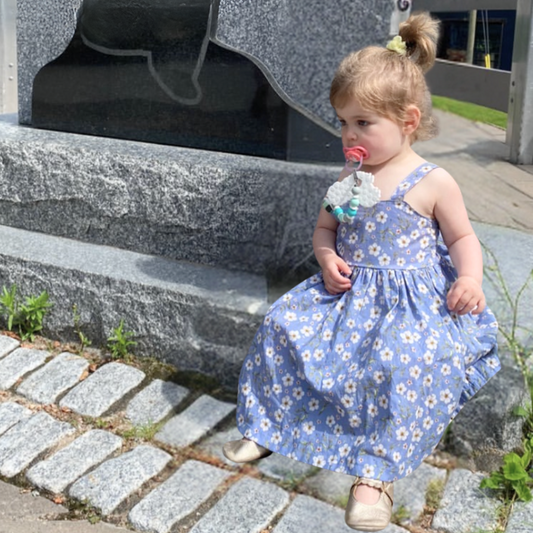 A toddler girl wearing a blue floral summer dress with an elasticized back, standing on a stone surface.