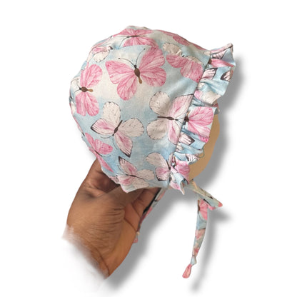 A handmade baby ruffle bonnet in baby blue with pink and white butterfly print, on a mannequin head held by a hand on a white background