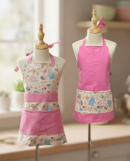 Two reversible handmade kids aprons displayed on child-sized dress forms in a bright kitchen setting. One apron features a full baking-themed print with pink pockets and straps, while the other shows the solid pink side with a coordinating printed pocket. Soft pastel lighting highlights the whimsical, child-friendly design.