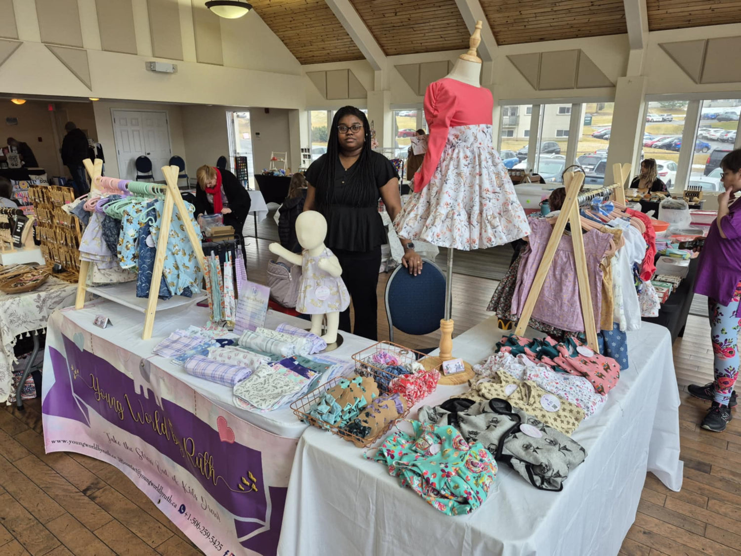 Ruth, founder of Young World by Ruth Inc., standing at her market booth showcasing handmade children’s clothing and accessories.