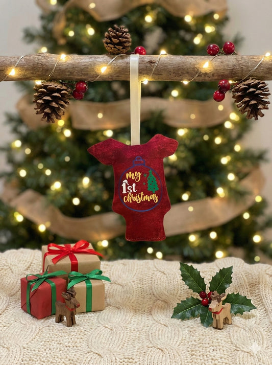 Mini red onesie ornament reading ‘My 1st Christmas’ hanging on a branch with pine cones and berries against a Christmas tree background surrounded by twinkly lights.