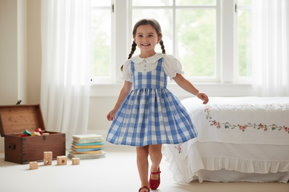 Young girl in a blue checkered dress standing in a bright room with a bed and wooden chest.